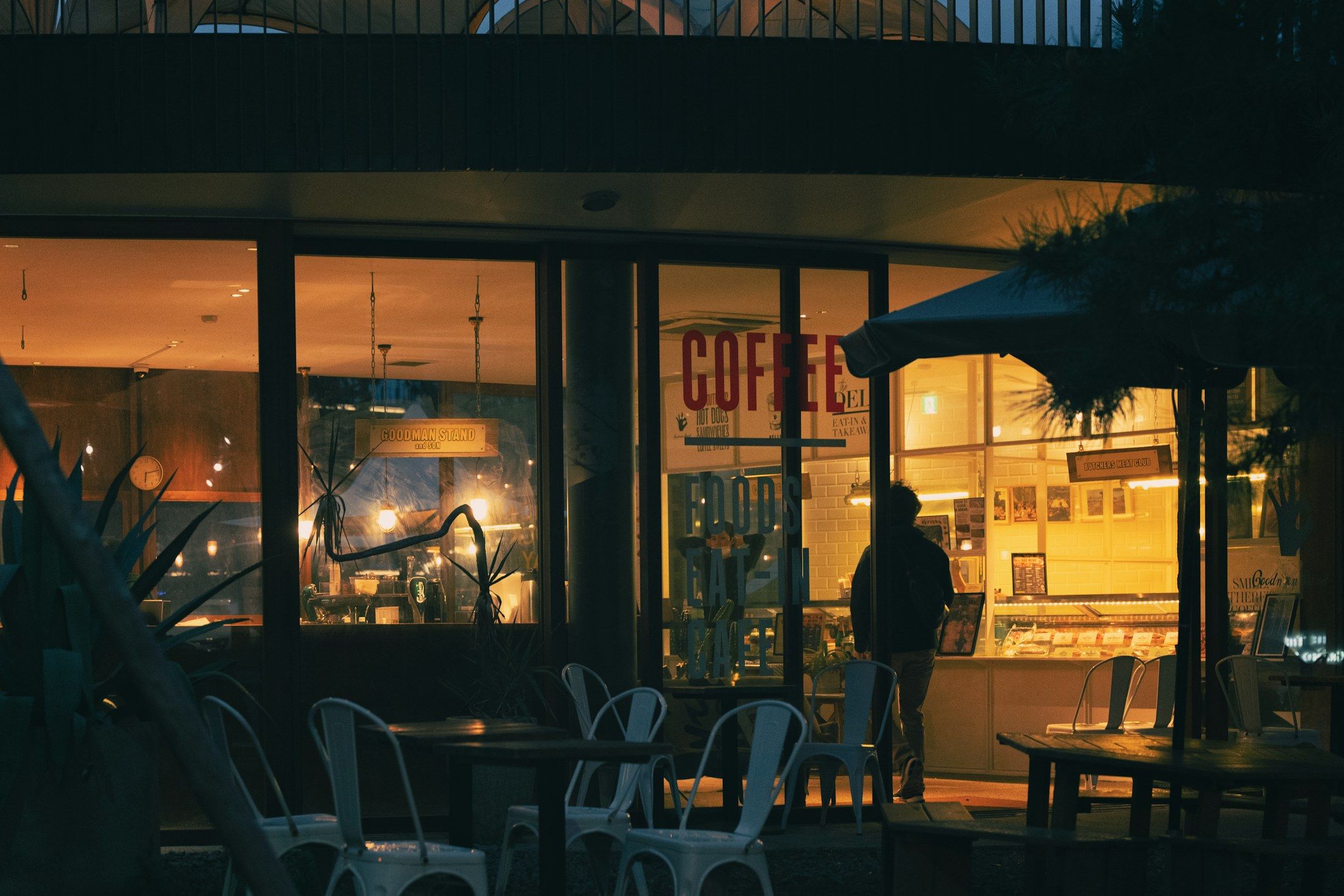 A coffee shop at dusk, warm light glowing from the windows onto the empty patio tables out front