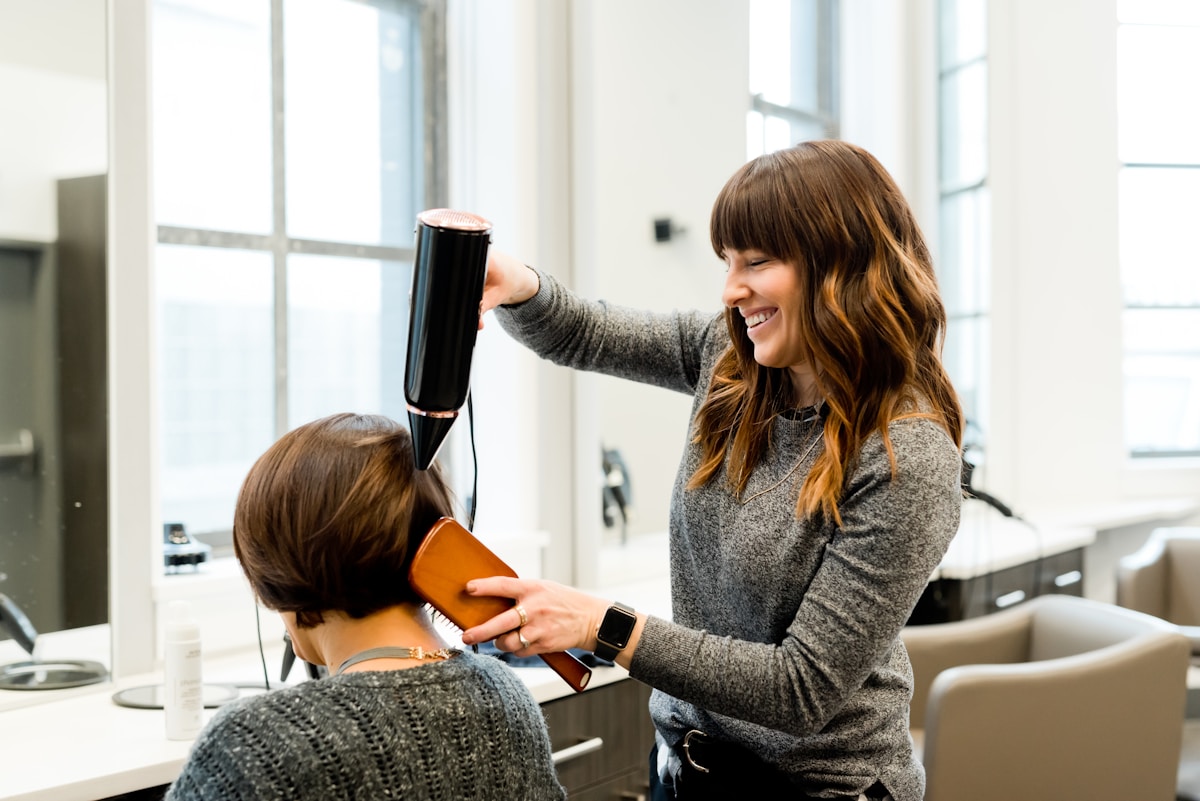 A stylist blow-drying a client's hair in the salon, big windows and soft daylight behind.