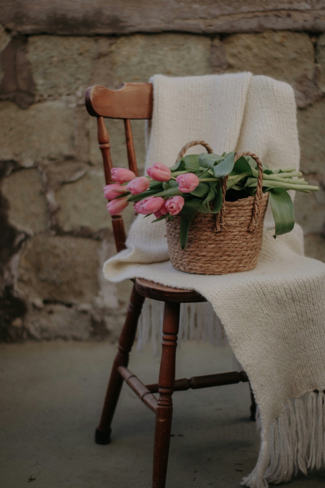 A handmade oat-colored blanket draped over a wooden chair beside a basket of tulips against a stone wall