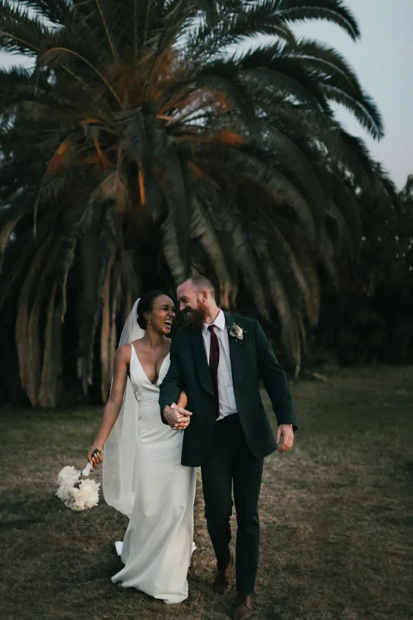 A couple walking together outdoors, holding hands, with trees behind them